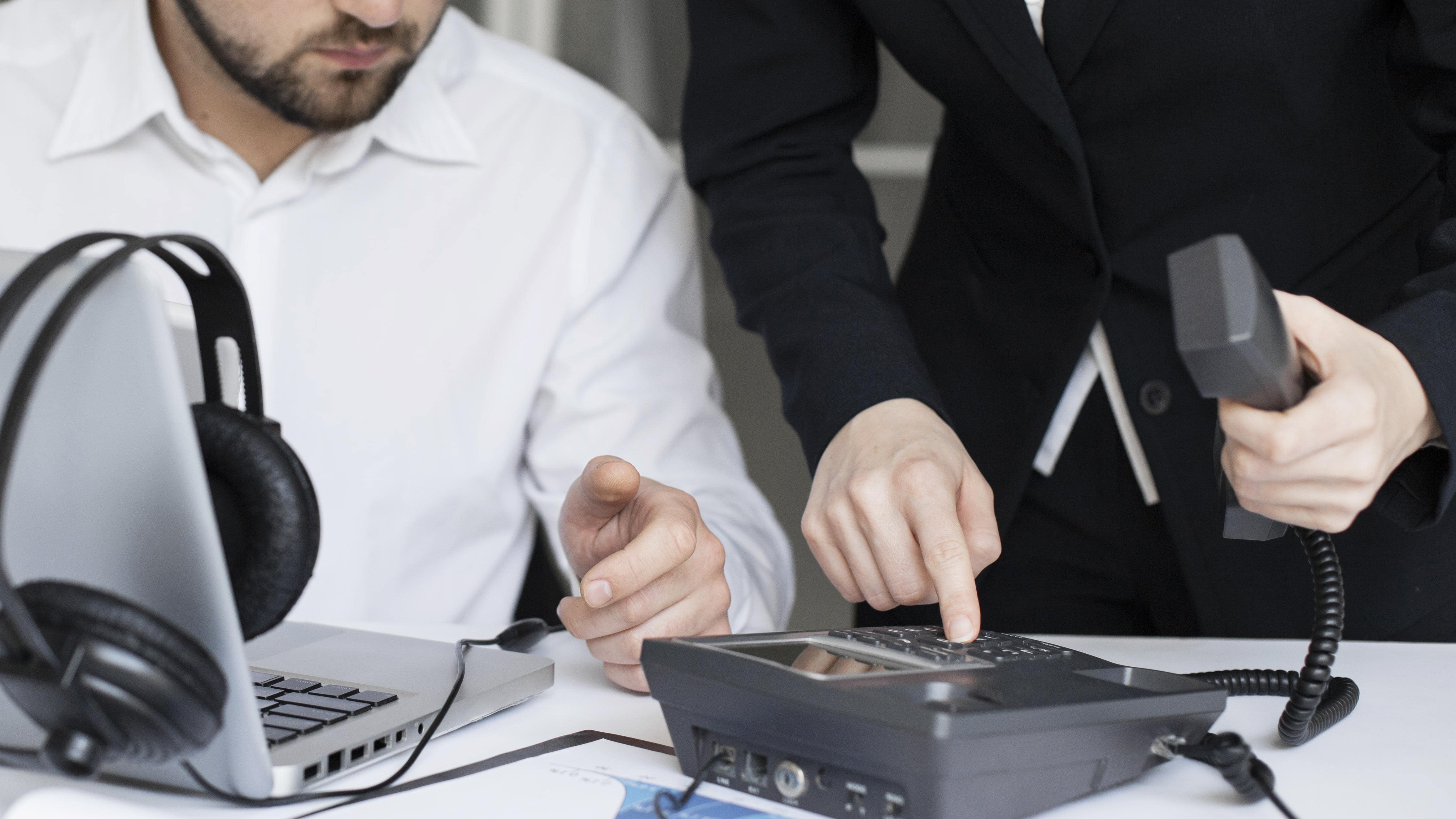 Close-up of two business people configuring a VoIP phone beside a laptop and headset, highlighting a hybrid communication setup in a modern workplace.